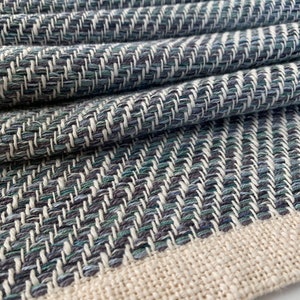 May include: A close-up of a woven fabric with a herringbone pattern in shades of blue, green, and white. The fabric has a natural, off-white border.