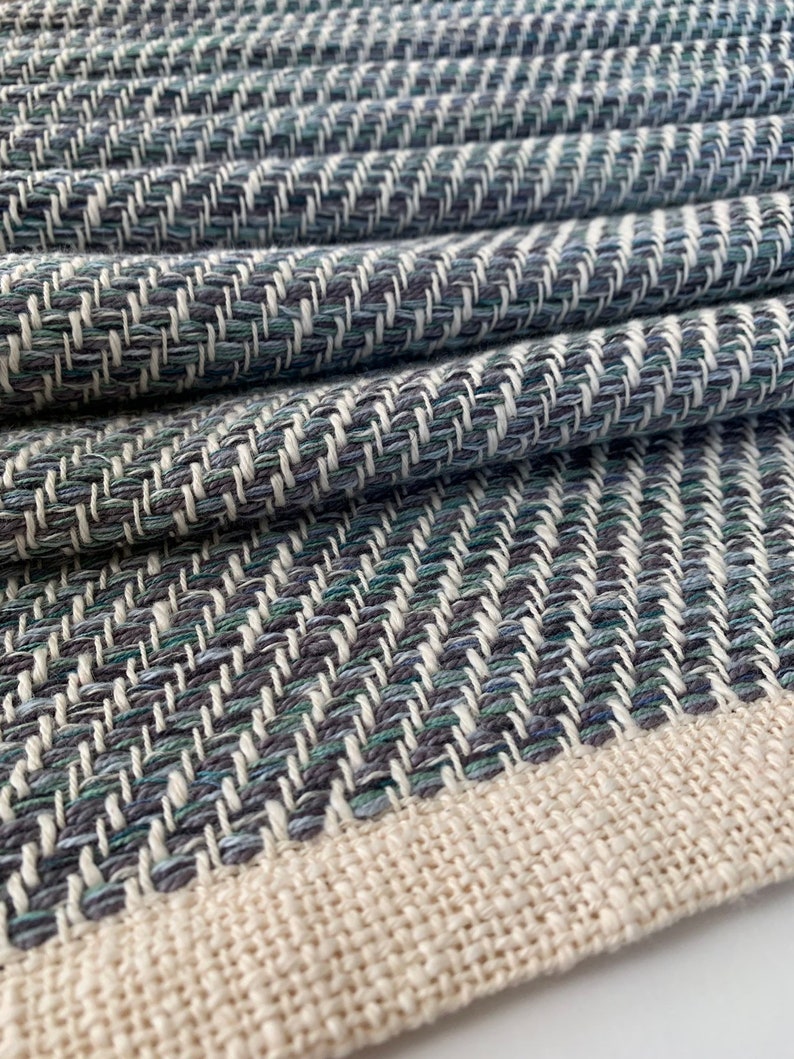 May include: A close-up of a woven fabric with a herringbone pattern in shades of blue, green, and white. The fabric has a natural, off-white border.