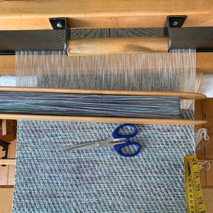 May include: A loom with a woven rug on the bottom. The rug is blue, green, and white with a herringbone pattern. A pair of scissors and a measuring tape are on the rug.