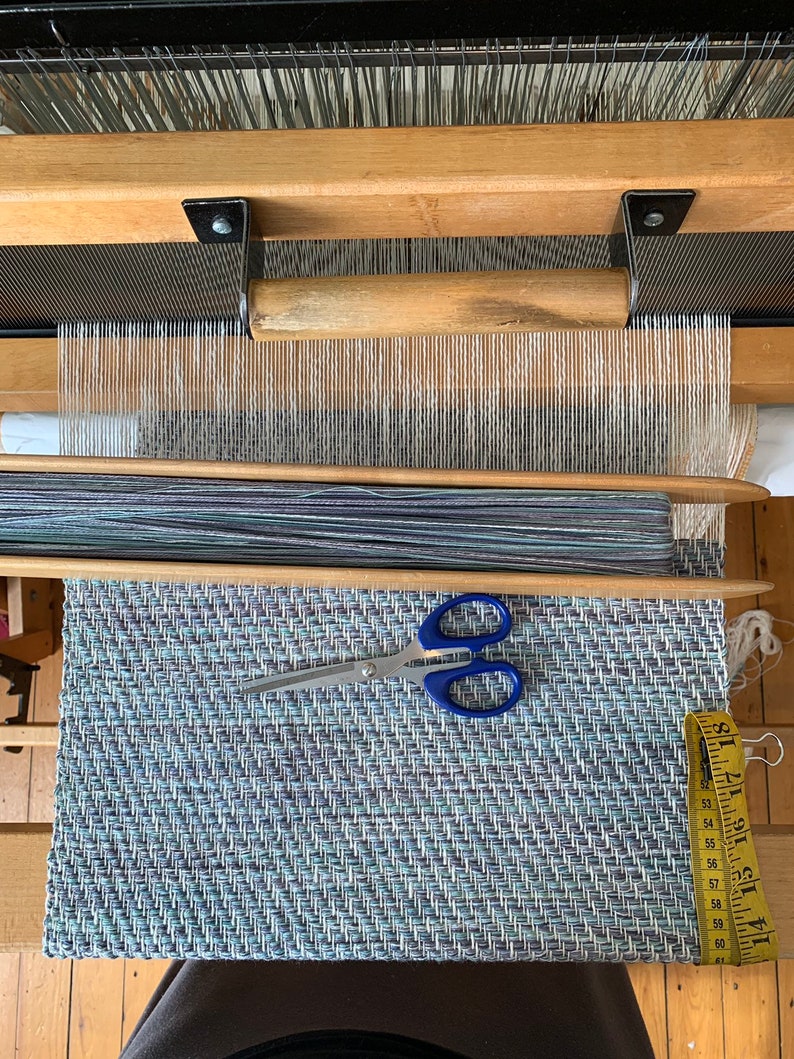 May include: A loom with a woven rug on the bottom. The rug is blue, green, and white with a herringbone pattern. A pair of scissors and a measuring tape are on the rug.