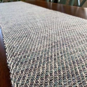 May include: A woven table runner in shades of gray, green, and white. The runner has a herringbone pattern and is made of a natural fiber.