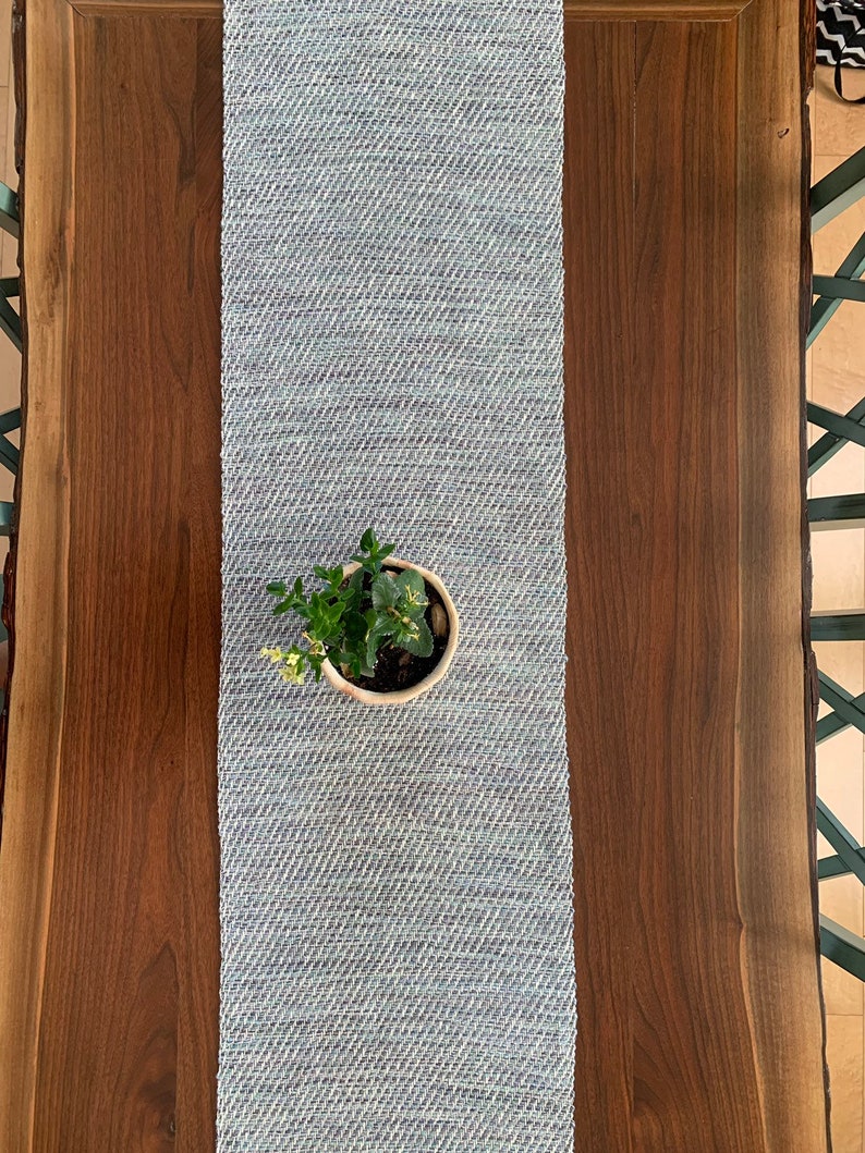 May include: A light blue woven table runner with a small potted plant in the center, on a dark brown wooden table.