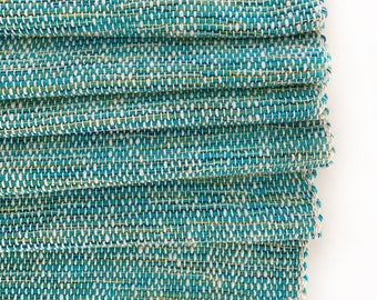 May include: Close-up of a stack of woven fabric in shades of teal, white, and green. The fabric has a textured appearance due to the weave. The edges of the fabric are visible, showing the layers. The background is plain white.