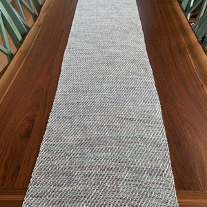 May include: A woven table runner in shades of blue, white, and purple. The runner has a subtle herringbone pattern and is laid out on a wooden table.