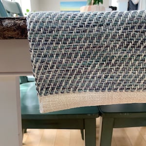 May include: A blue and white woven table runner with a chevron pattern. The runner has a white border and is hanging over the edge of a table.