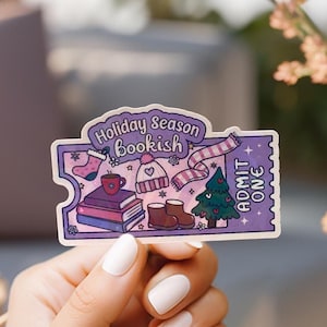 May include: A purple sticker with the text "Holiday Season bookish" and illustrations of winter items like a hat, scarf, boots, and a Christmas tree. The sticker is held by a person with white nail polish.