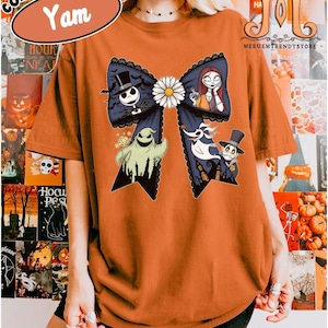 Peut inclure: T-shirt orange Comfort Colors avec un grand n&oelig;ud bleu orn&eacute; de personnages de L'&Eacute;trange No&euml;l de Monsieur Jack. Le n&oelig;ud a une marguerite blanche au centre et une bordure en dentelle noire. Le t-shirt porte &eacute;galement le mot "Yam" dans un ovale blanc.