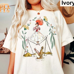 Puede incluir: Una camiseta blanca con un estampado gráfico de un personaje de dibujos animados sosteniendo un paraguas, de pie sobre una cuerda floja sobre un cocodrilo. El personaje lleva un vestido rojo y un sombrero blanco. El fondo es un diseño floral verde y amarillo.