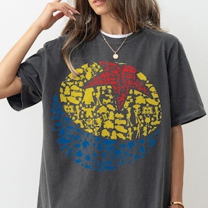 May include: Dark gray t-shirt featuring a large graphic of a red star inside a yellow circle, with blue accents. The graphic is filled with various shapes and figures. The shirt has short sleeves and a relaxed fit.