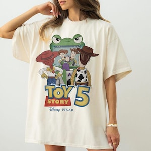 May include: Cream-colored t-shirt featuring a graphic from the movie "Toy Story 5". The design includes cartoon characters, the movie title, and the Disney Pixar logo. The shirt has short sleeves and a relaxed fit.