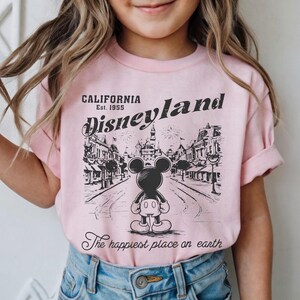 以下が含まれることがあります： ピンク色のTシャツ。ミッキーマウスがディズニーランドを背景にした白黒のグラフィック。テキストには「CALIFORNIA Est. 1955 Disneyland」と「The happiest place on earth」が含まれています。