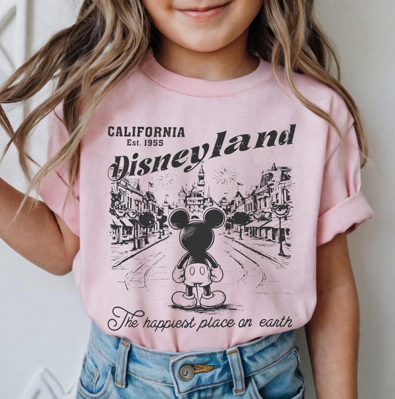 以下が含まれることがあります： ピンク色のTシャツ。ミッキーマウスがディズニーランドを背景にした白黒のグラフィック。テキストには「CALIFORNIA Est. 1955 Disneyland」と「The happiest place on earth」が含まれています。