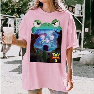 May include: A pale pink t-shirt with a Toy Story 5 graphic. The graphic features Woody, Buzz Lightyear, and Jessie. The design is framed by a green frog shape. The shirt is a casual, oversized style.
