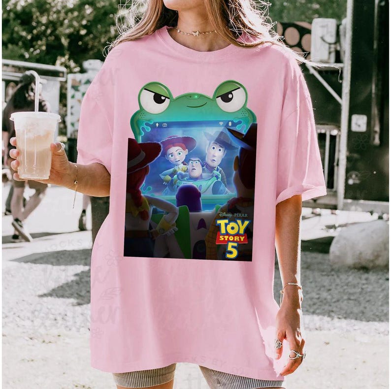 May include: A pale pink t-shirt with a Toy Story 5 graphic. The graphic features Woody, Buzz Lightyear, and Jessie. The design is framed by a green frog shape. The shirt is a casual, oversized style.