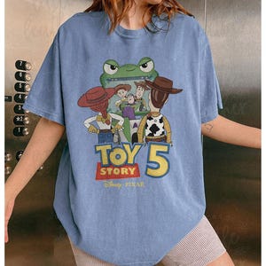 May include: A light blue t-shirt featuring a graphic from the movie Toy Story 5. The graphic includes characters like Woody, Buzz Lightyear, and Jessie, with the text "TOY STORY 5" in a colorful design. The Disney Pixar logo is at the bottom.