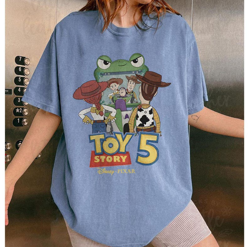 May include: A light blue t-shirt featuring a graphic from the movie Toy Story 5. The graphic includes characters like Woody, Buzz Lightyear, and Jessie, with the text "TOY STORY 5" in a colorful design. The Disney Pixar logo is at the bottom.