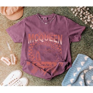 May include: A purple t-shirt featuring the text "Lightning McQueen" and a graphic of a race car. The shirt is styled with white sneakers, a corduroy hat, and a blue blanket with white hearts. A delicate necklace is also visible.