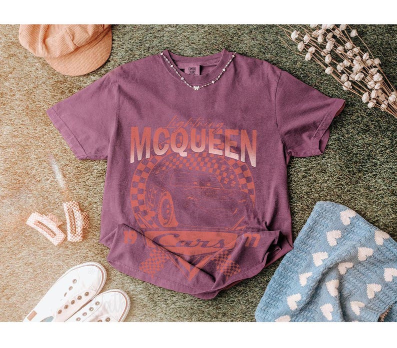 May include: A purple t-shirt featuring the text "Lightning McQueen" and a graphic of a race car. The shirt is styled with white sneakers, a corduroy hat, and a blue blanket with white hearts. A delicate necklace is also visible.