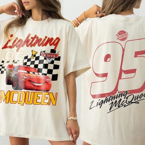 May include: Cream-colored t-shirt featuring a graphic of Lightning McQueen from the Cars movie. The front design includes the text "Lightning McQueen 95" with a checkered flag and trophy. The back displays a large "95" and "Lightning McQueen" text.