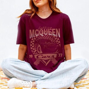 May include: Burgundy t-shirt featuring a graphic of Lightning McQueen from the movie Cars. The design includes the text "Lightning McQueen" above a checkered circle with the car's image, and "19 Cars 77" below. The shirt is worn by a person.
