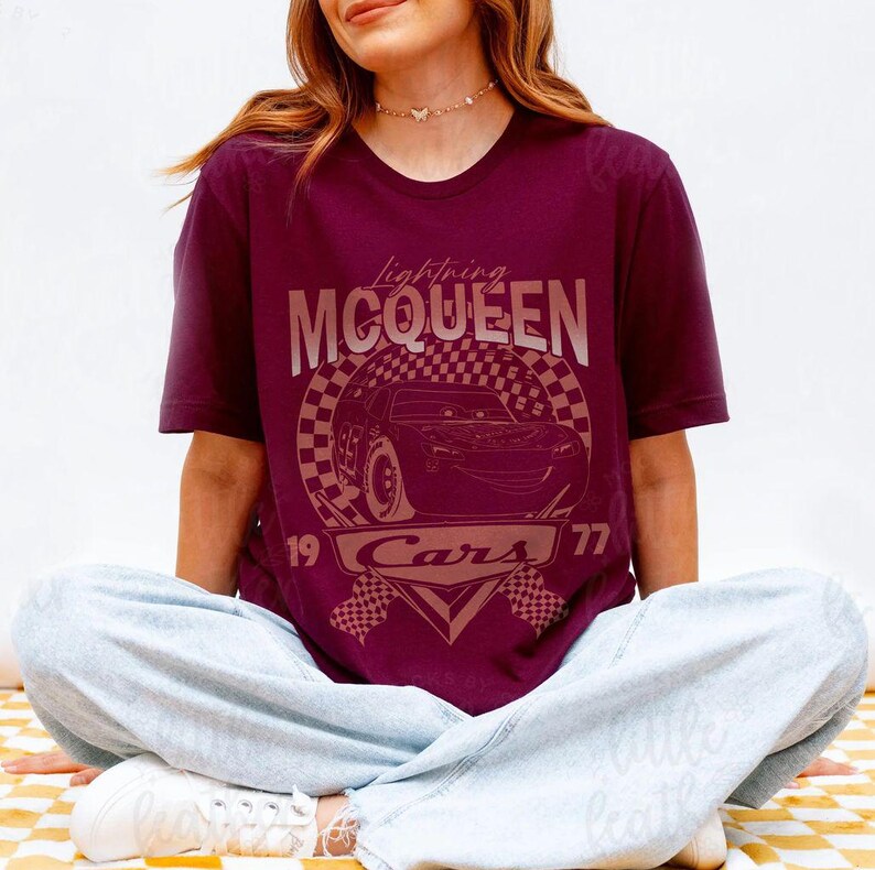 May include: Burgundy t-shirt featuring a graphic of Lightning McQueen from the movie Cars. The design includes the text "Lightning McQueen" above a checkered circle with the car's image, and "19 Cars 77" below. The shirt is worn by a person.