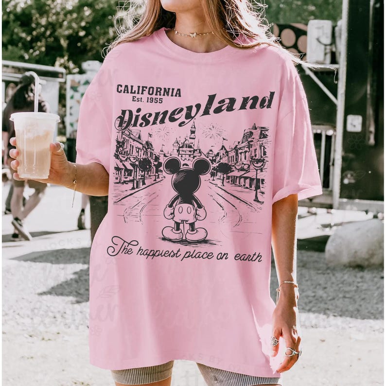 以下が含まれることがあります： ピンク色のTシャツ。ディズニーランドのグラフィックがプリントされています。ミッキーマウスが街並みを背景に描かれ、「California Est. 1955 Disneyland」と「The happiest place on earth」の文字が入っています。