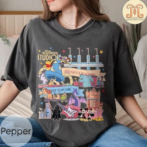 Può includere: Una maglietta grigia con una grafica colorata di vari personaggi e attrazioni Disney dei Disney's Hollywood Studios. La grafica include Topolino, Minnie Mouse, Woody, Buzz Lightyear, Darth Vader e altro ancora. Il testo "Disney's Hollywood Studios" e "Hollywood Boulevard" sono anche inclusi nella grafica.