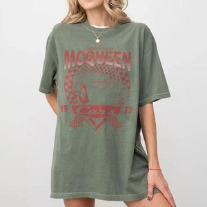 May include: An olive green t-shirt featuring a red graphic of Lightning McQueen from the Cars movie. The design includes the text "Lightning McQueen", "Cars", and the numbers "19" and "77". The shirt has short sleeves and a crew neck.