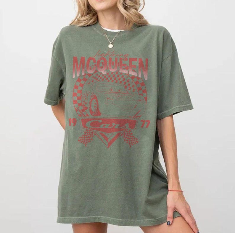 May include: An olive green t-shirt featuring a red graphic of Lightning McQueen from the Cars movie. The design includes the text "Lightning McQueen", "Cars", and the numbers "19" and "77". The shirt has short sleeves and a crew neck.