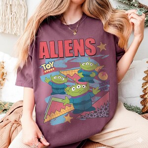 May include: A maroon t-shirt featuring the word "ALIENS" in red block letters with a retro design. The shirt has a graphic of three green aliens from Toy Story, with the text "TOY STORY" and "SINCE 1995".