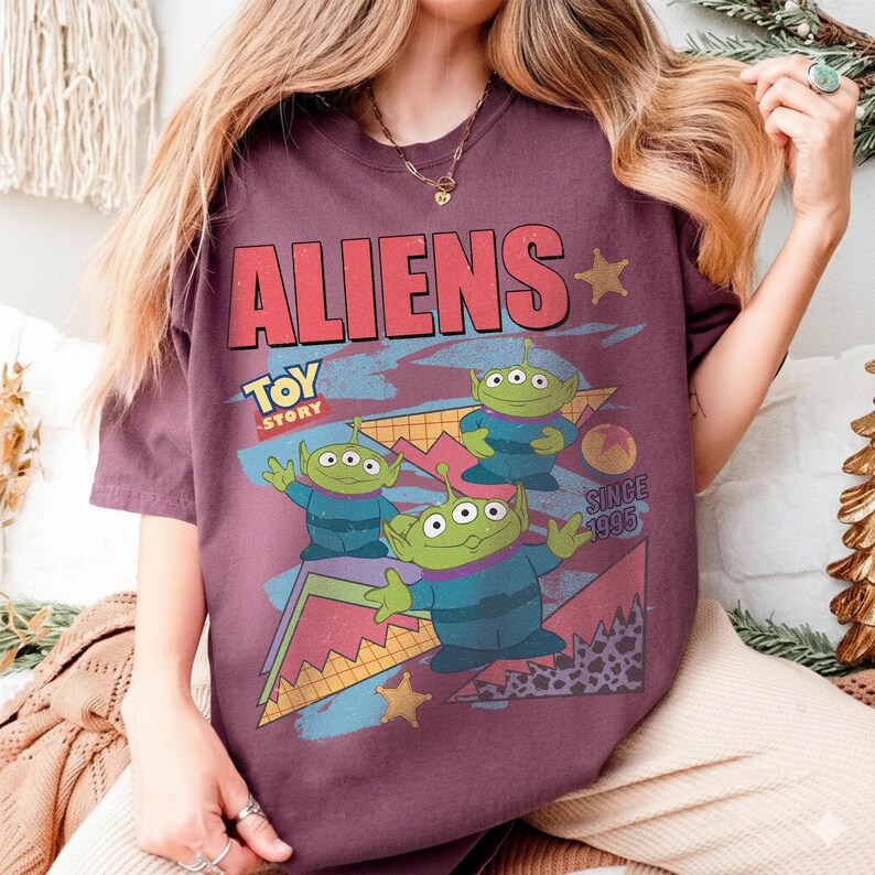 May include: A maroon t-shirt featuring the word "ALIENS" in red block letters with a retro design. The shirt has a graphic of three green aliens from Toy Story, with the text "TOY STORY" and "SINCE 1995".