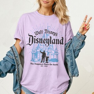 Puede incluir: Camiseta lila claro con la inscripci&oacute;n "Walt Disney's Disneyland" en negro y un gr&aacute;fico azul de un castillo. Debajo, el texto "The Happiest Place On Earth EST. 1955". La camiseta tiene mangas cortas y cuello redondo.