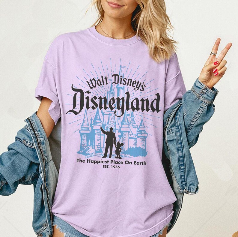 Puede incluir: Camiseta lila claro con la inscripci&oacute;n "Walt Disney's Disneyland" en negro y un gr&aacute;fico azul de un castillo. Debajo, el texto "The Happiest Place On Earth EST. 1955". La camiseta tiene mangas cortas y cuello redondo.