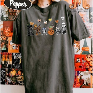 Op de afbeelding: Donkergrijs Comfort Colors T-shirt met een Halloween-thema ontwerp. Het ontwerp bevat cartoonfiguren met ballonvormige hoofden. Het shirt heeft het woord "Pepper" in een witte ovaal. De achtergrond bevat Halloween-posters.
