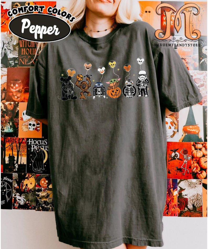 Op de afbeelding: Donkergrijs Comfort Colors T-shirt met een Halloween-thema ontwerp. Het ontwerp bevat cartoonfiguren met ballonvormige hoofden. Het shirt heeft het woord "Pepper" in een witte ovaal. De achtergrond bevat Halloween-posters.