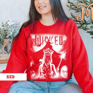 May include: A red sweatshirt featuring a white graphic of the word "WICKED" with a silhouette of a witch and a woman with a crown and wand. The word "RED" is printed on a white rectangle at the bottom.