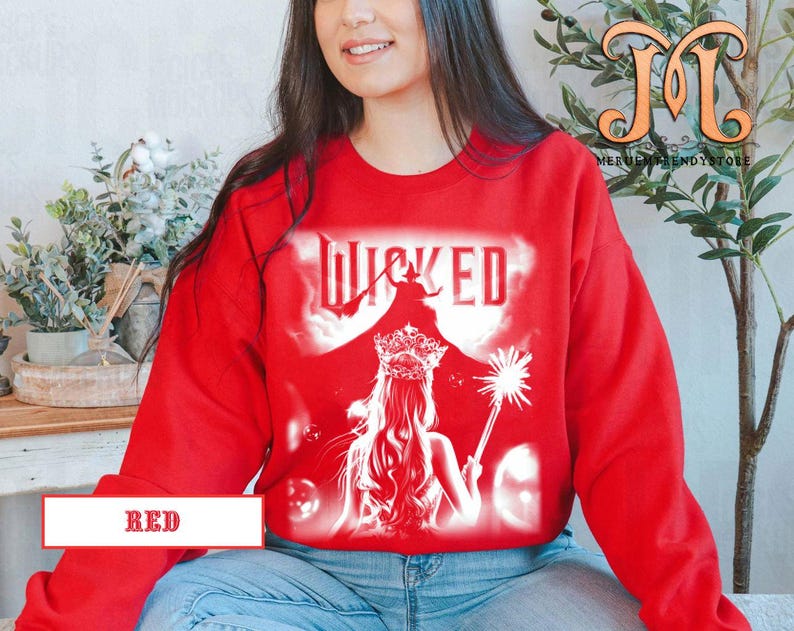May include: A red sweatshirt featuring a white graphic of the word "WICKED" with a silhouette of a witch and a woman with a crown and wand. The word "RED" is printed on a white rectangle at the bottom.