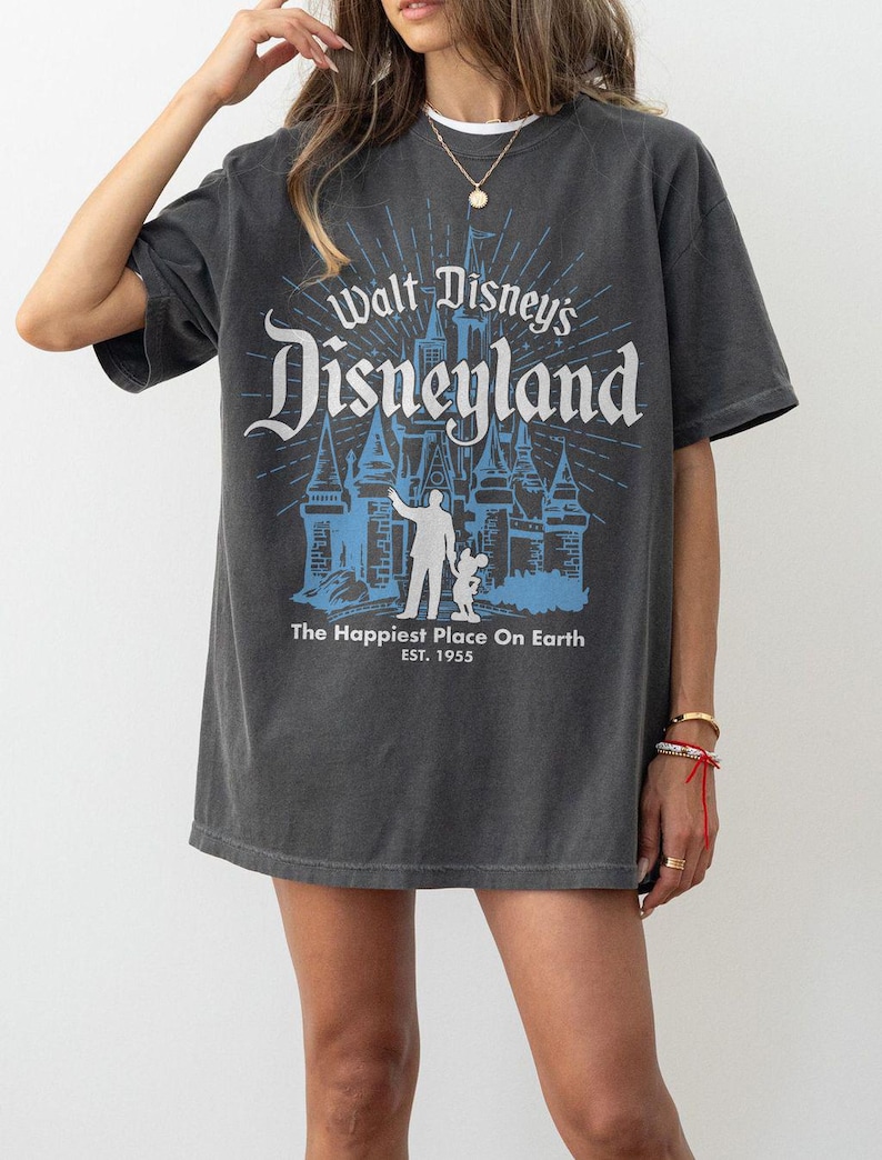 Puede incluir: Camiseta gris oscuro con un dise&ntilde;o azul y blanco de Disneyland. El dise&ntilde;o incluye el castillo de Disneyland, el texto "Walt Disney's Disneyland" y "The Happiest Place On Earth EST. 1955". La camiseta tiene mangas cortas.