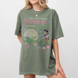 May include: Olive green t-shirt featuring the text "EPCOT INTERNATIONAL FLOWER & GARDEN FESTIVAL" with a graphic of a floral sphere, Mickey Mouse, and butterflies. The shirt has short sleeves and a relaxed fit.