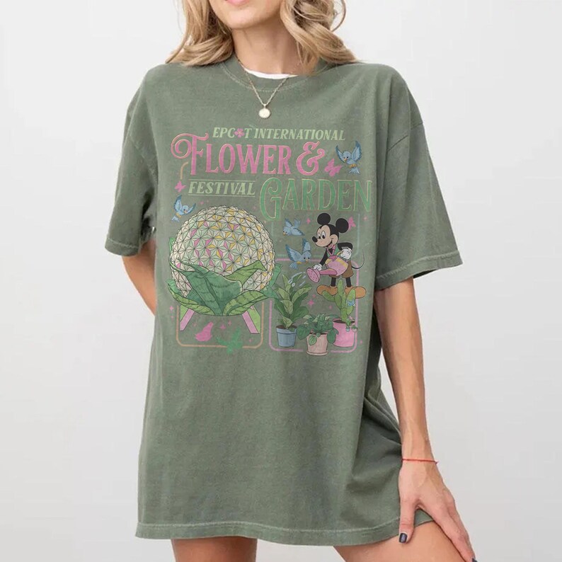 May include: Olive green t-shirt featuring the text "EPCOT INTERNATIONAL FLOWER & GARDEN FESTIVAL" with a graphic of a floral sphere, Mickey Mouse, and butterflies. The shirt has short sleeves and a relaxed fit.