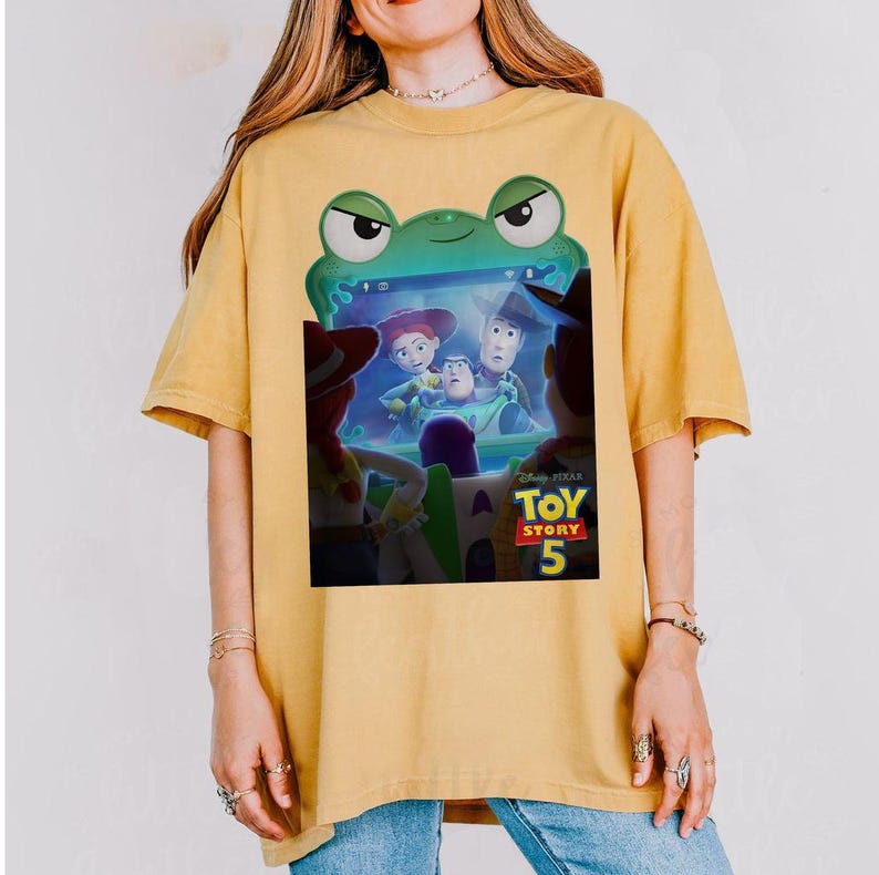 May include: Mustard yellow t-shirt with a graphic from the film Toy Story 5. The design features Woody, Buzz Lightyear, Jessie, and other characters within a green frog-shaped frame. The Disney Pixar logo is also present.