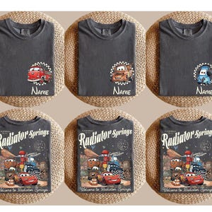 Op de afbeelding: Zes antracietgrijze t-shirts. De bovenste rij heeft drie shirts met cartoon autografische afbeeldingen en de tekst "Name". De onderste rij toont drie shirts met een "Radiator Springs" ontwerp en de tekst "Welcome to Radiator Springs".