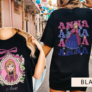 May include: Two black t-shirts with different Anna from Frozen designs. One shirt features a cartoon image of Anna in a pink frame with flowers and the word "Anna" below. The other shirt has a repeated image of Anna in a purple dress with the word "Anna" in a retro font.