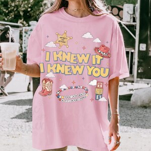 May include: A light pink t-shirt featuring the yellow text I KNEW IT I KNEW YOU. The graphic includes a sheriff star with the text INFINITY AND BEYOND, a red cowboy hat, a pair of red cowboy boots, a beaded bracelet with the text YOU'VE GOT A FRIEND IN ME, and a stack of alphabet blocks.