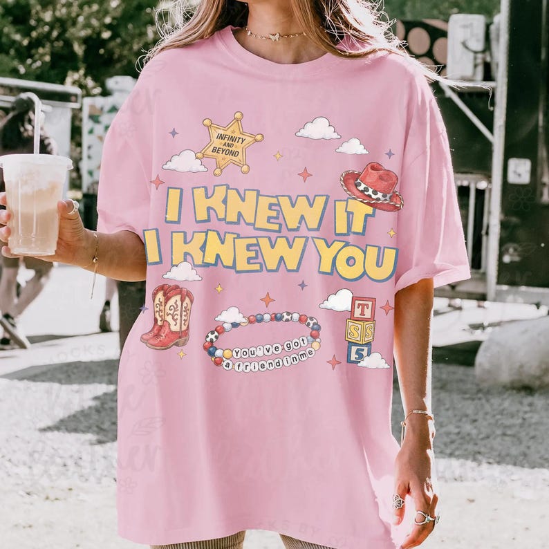 May include: A light pink t-shirt featuring the yellow text I KNEW IT I KNEW YOU. The graphic includes a sheriff star with the text INFINITY AND BEYOND, a red cowboy hat, a pair of red cowboy boots, a beaded bracelet with the text YOU'VE GOT A FRIEND IN ME, and a stack of alphabet blocks.