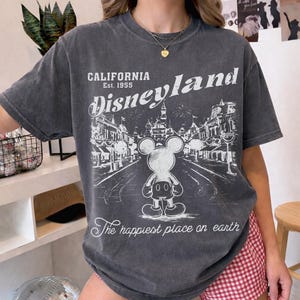 以下が含まれることがあります： ディズニーランドのグラフィックがプリントされた濃いグレーのTシャツ。「CALIFORNIA Est. 1955 Disneyland」の文字と、ミッキーマウスのイラストが街並みを背景に描かれています。「The happiest place on earth」の文字も含まれています。