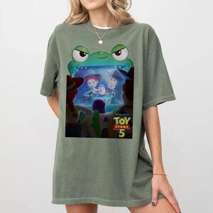 May include: Sage green t-shirt with a graphic from the film Toy Story 5. The graphic depicts characters from the film within a green frog-shaped frame. The t-shirt has a relaxed fit.