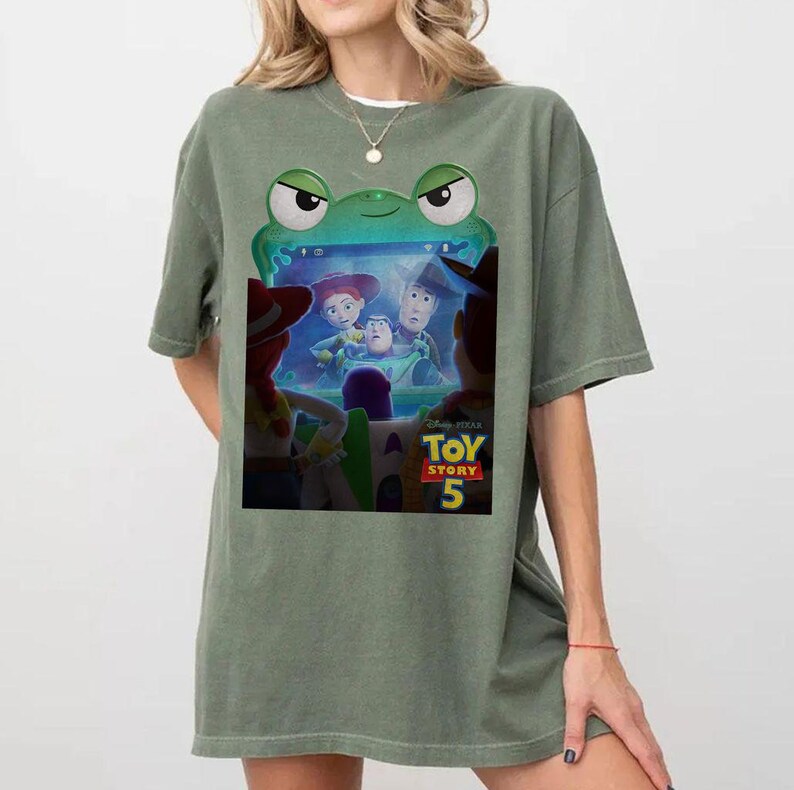 May include: Sage green t-shirt with a graphic from the film Toy Story 5. The graphic depicts characters from the film within a green frog-shaped frame. The t-shirt has a relaxed fit.