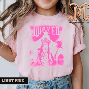 May include: Light pink t-shirt featuring a pink graphic of a woman wearing a crown and holding a wand, with the word "WICKED" above. The shirt is light pink with short sleeves. The words "LIGHT PINK" are in a black rectangle at the bottom.