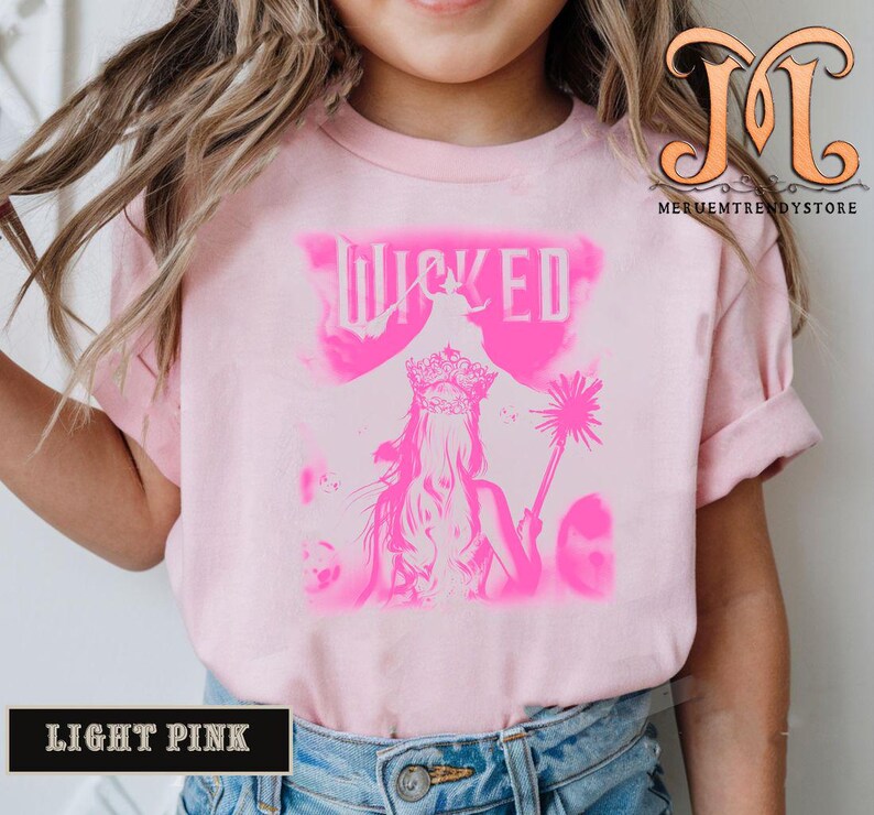 May include: Light pink t-shirt featuring a pink graphic of a woman wearing a crown and holding a wand, with the word "WICKED" above. The shirt is light pink with short sleeves. The words "LIGHT PINK" are in a black rectangle at the bottom.